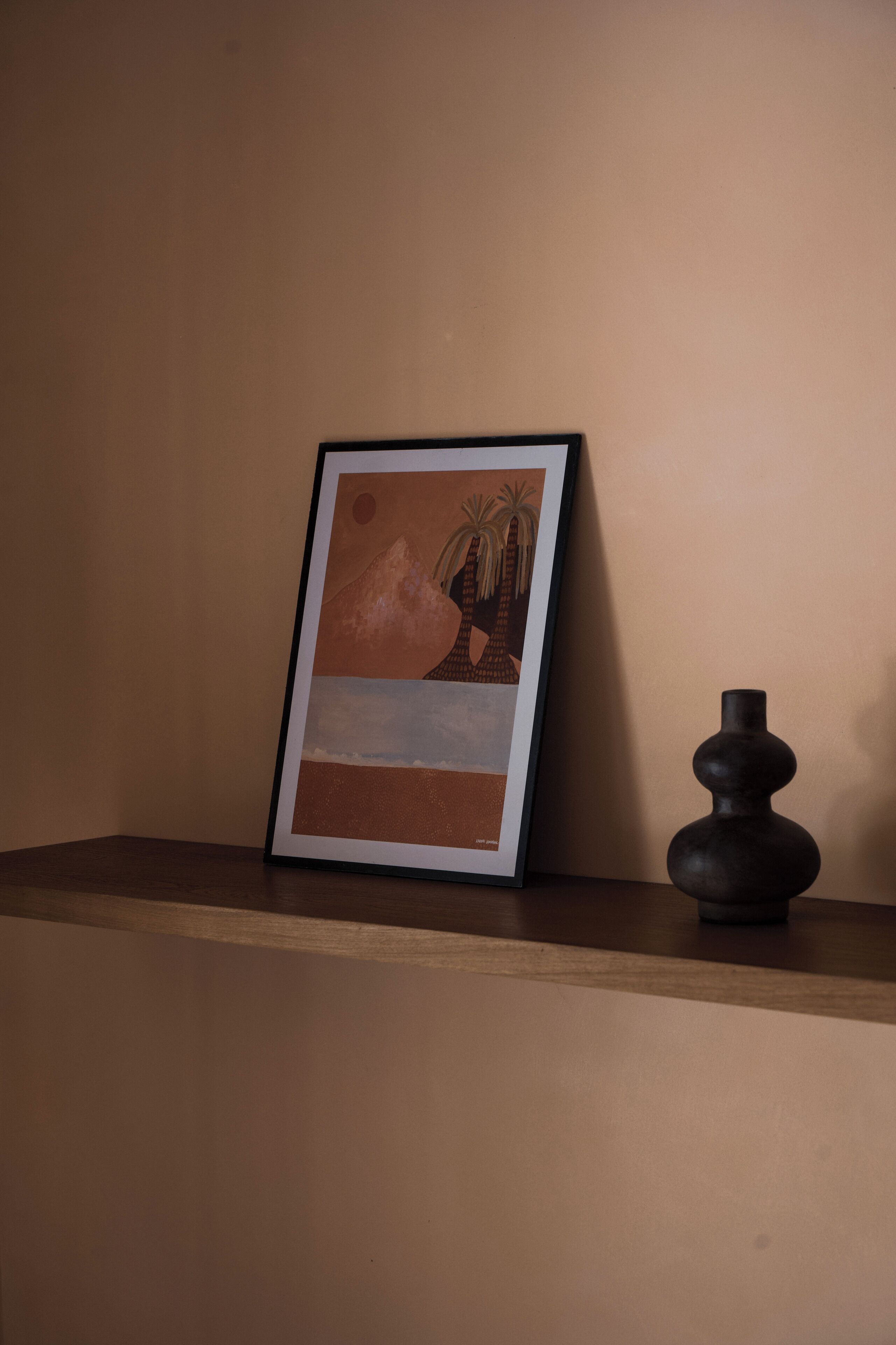 Una obra de arte enmarcada en una pared, un jarrón oscuro sobre una estantería y un repisa de madera.