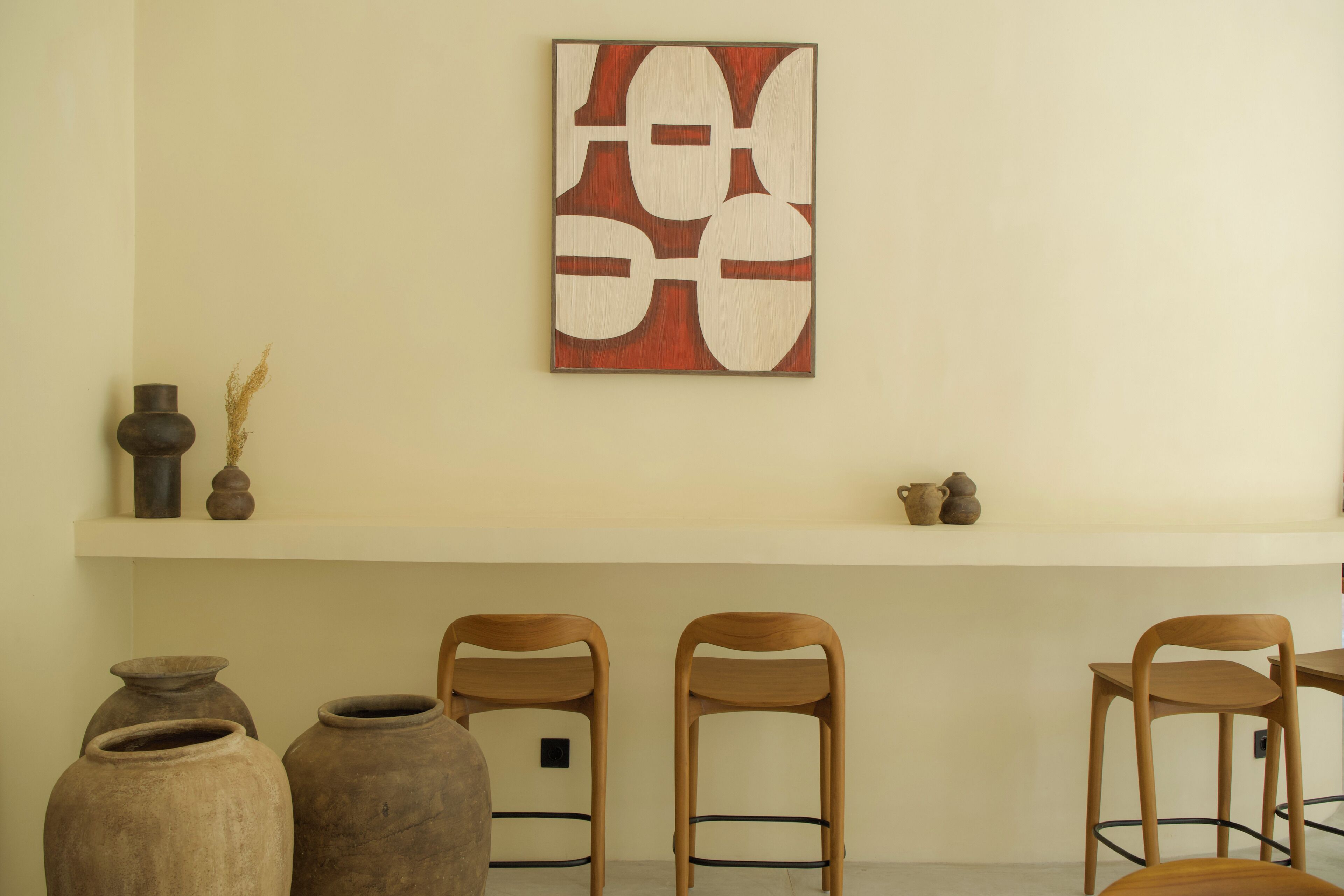 Una habitación con pared blanca, un estante de madera con cerámica y tres sillas de madera.