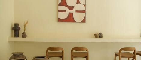 Una habitación con pared blanca, un estante de madera con cerámica y tres sillas de madera.