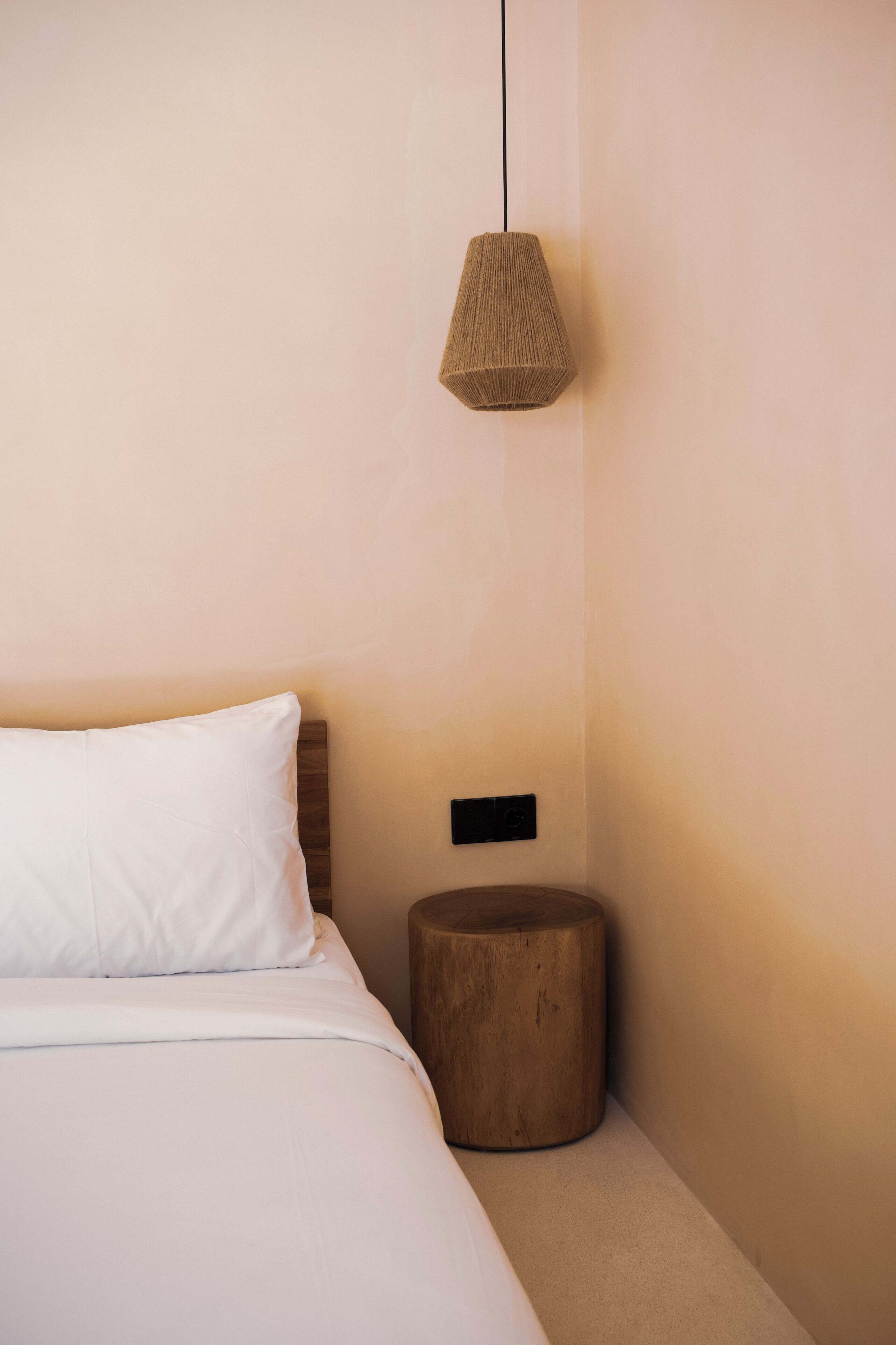 A bed with white bedding, a round wooden nightstand, and a woven pendant light.