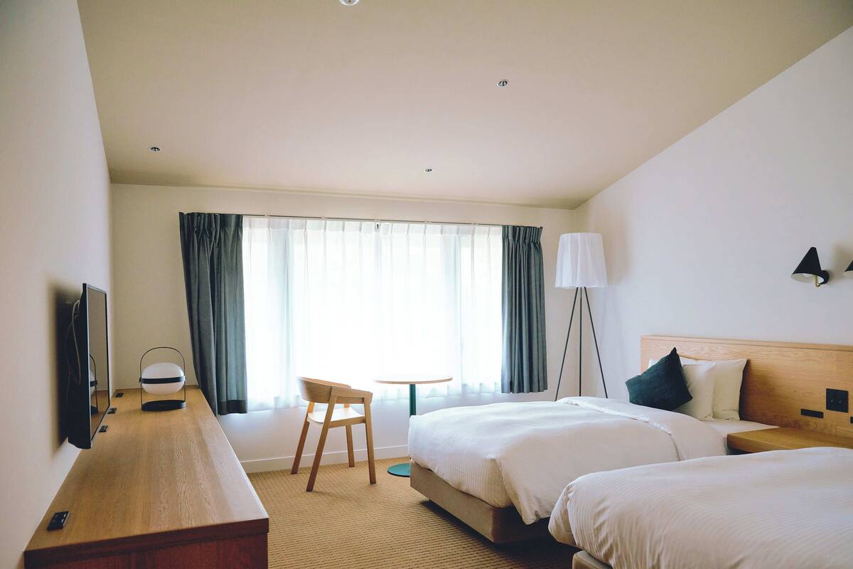 Hotellihuone, jossa on kaksi sänkyä, työpöytä tuolilla, lattiavalaisin ja suuri ikkuna, jossa on verhot.
