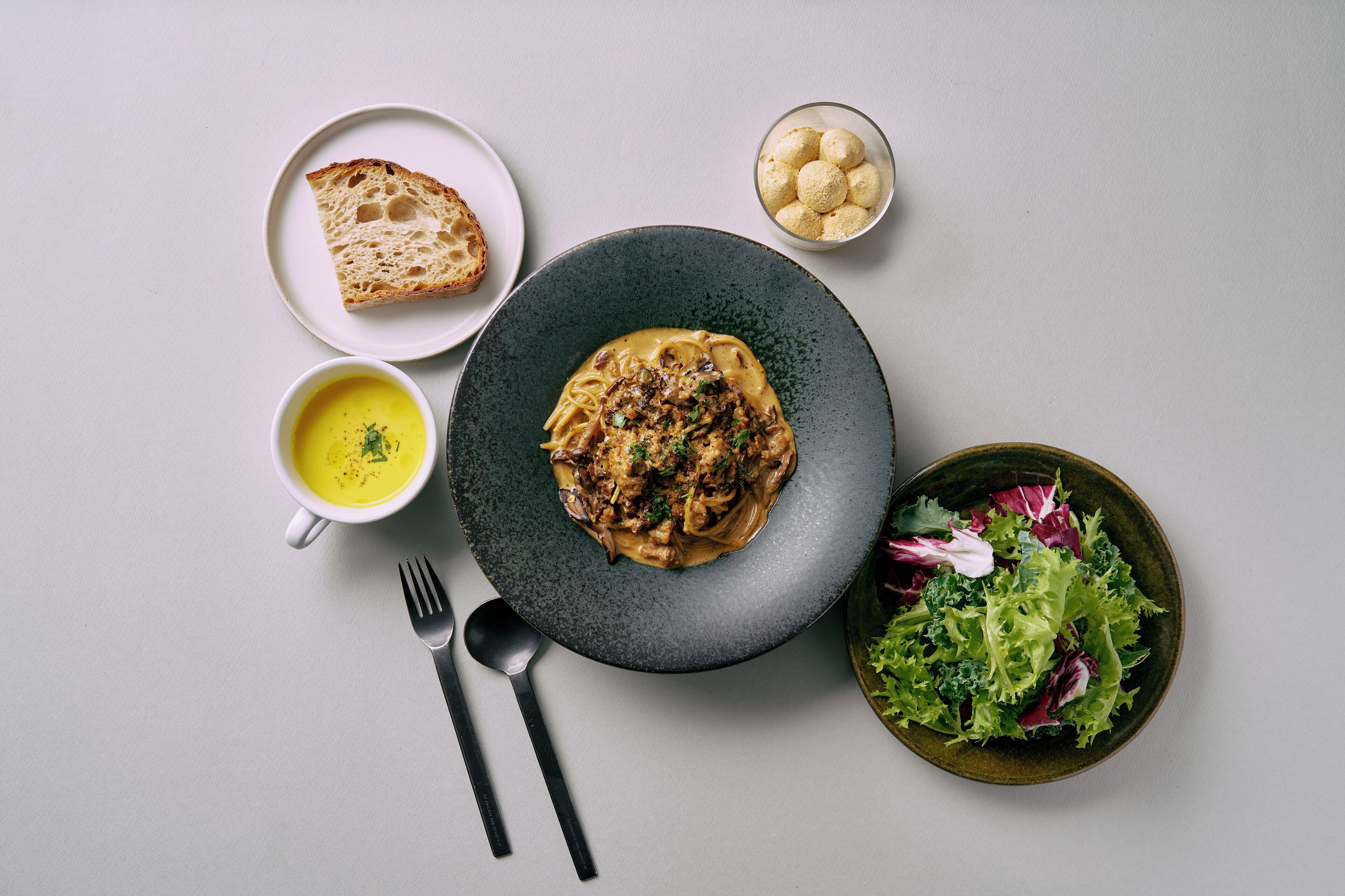 A plate of pasta with a creamy sauce, a bowl of soup, a plate of bread, a bowl of salad, and a small bowl of round bread-like items.