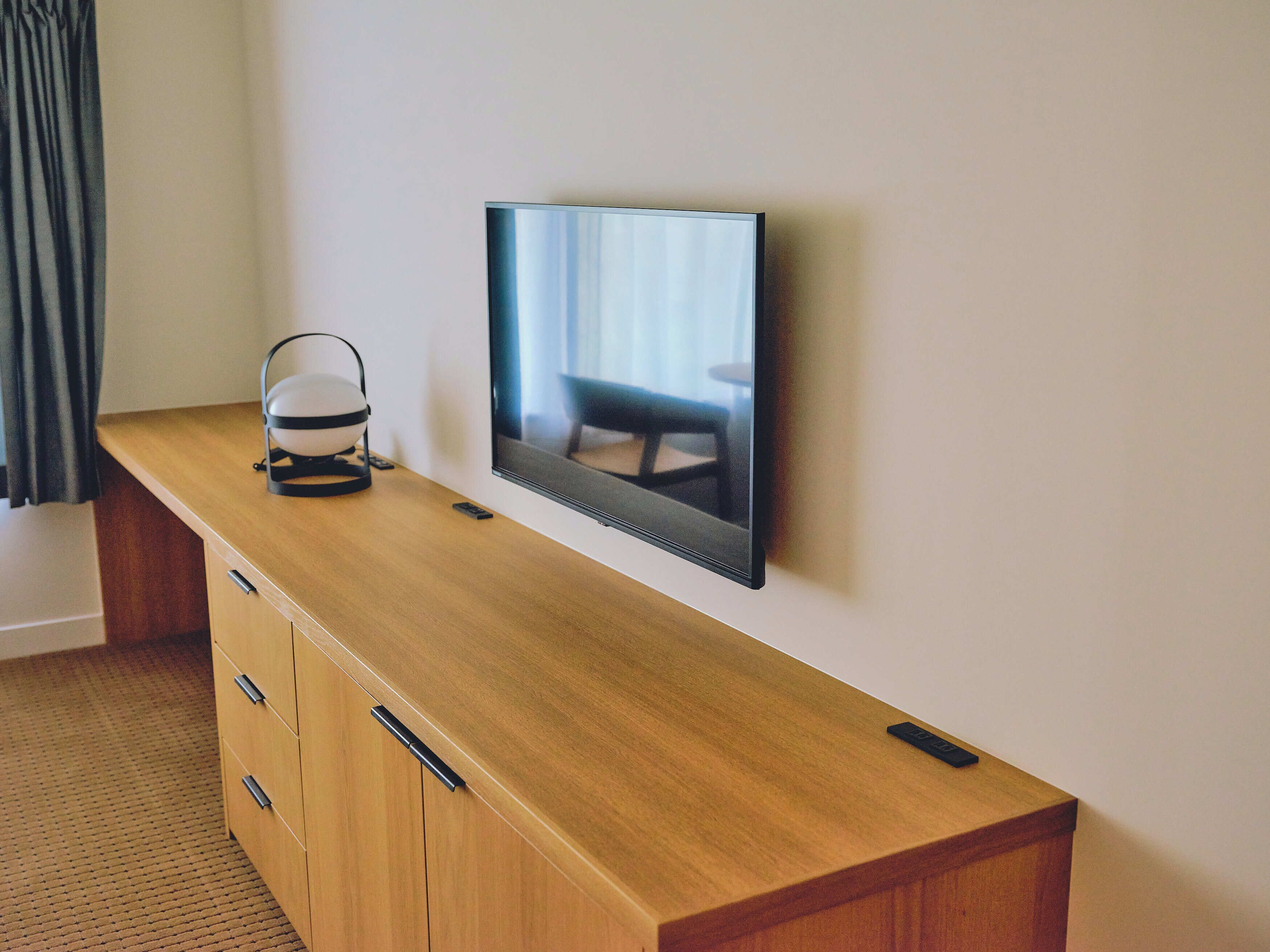A modern room with a wooden desk, a flat-screen TV mounted on the wall, and a small bedside table with a lamp.