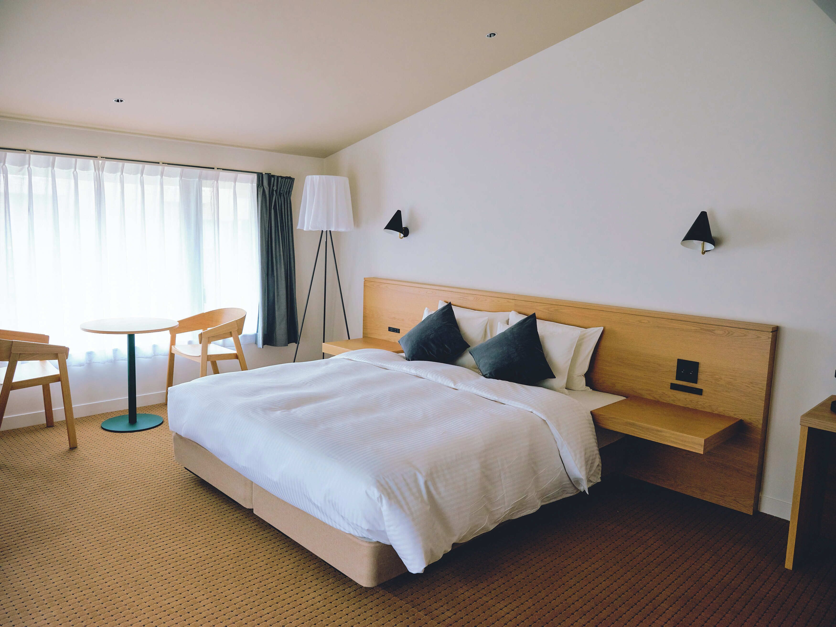 A hotel room with a large bed, a small table, two chairs, a floor lamp, and a window with curtains.