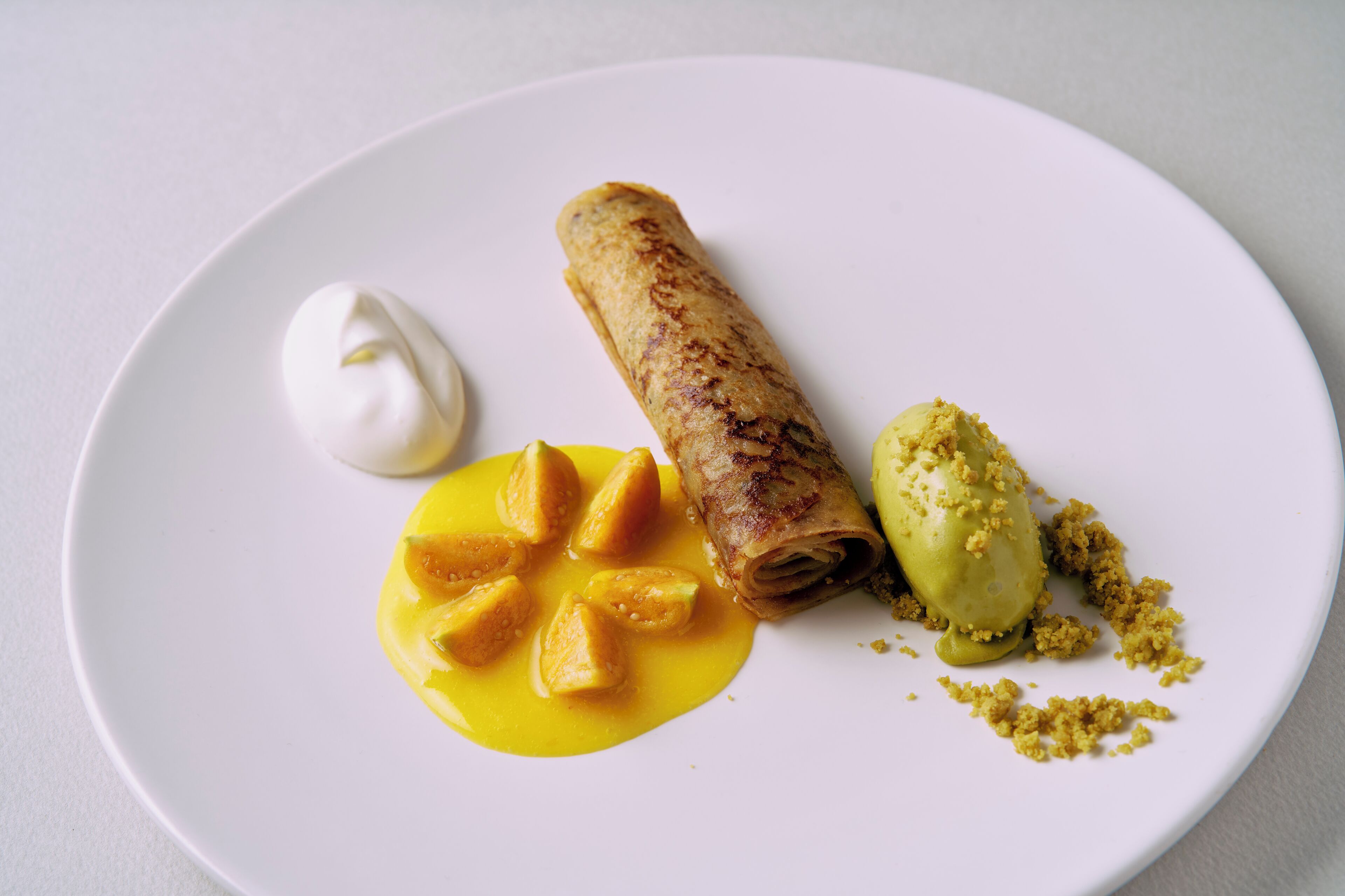A plate with a rolled item, a dollop of cream, and a yellow sauce with cubed food.
