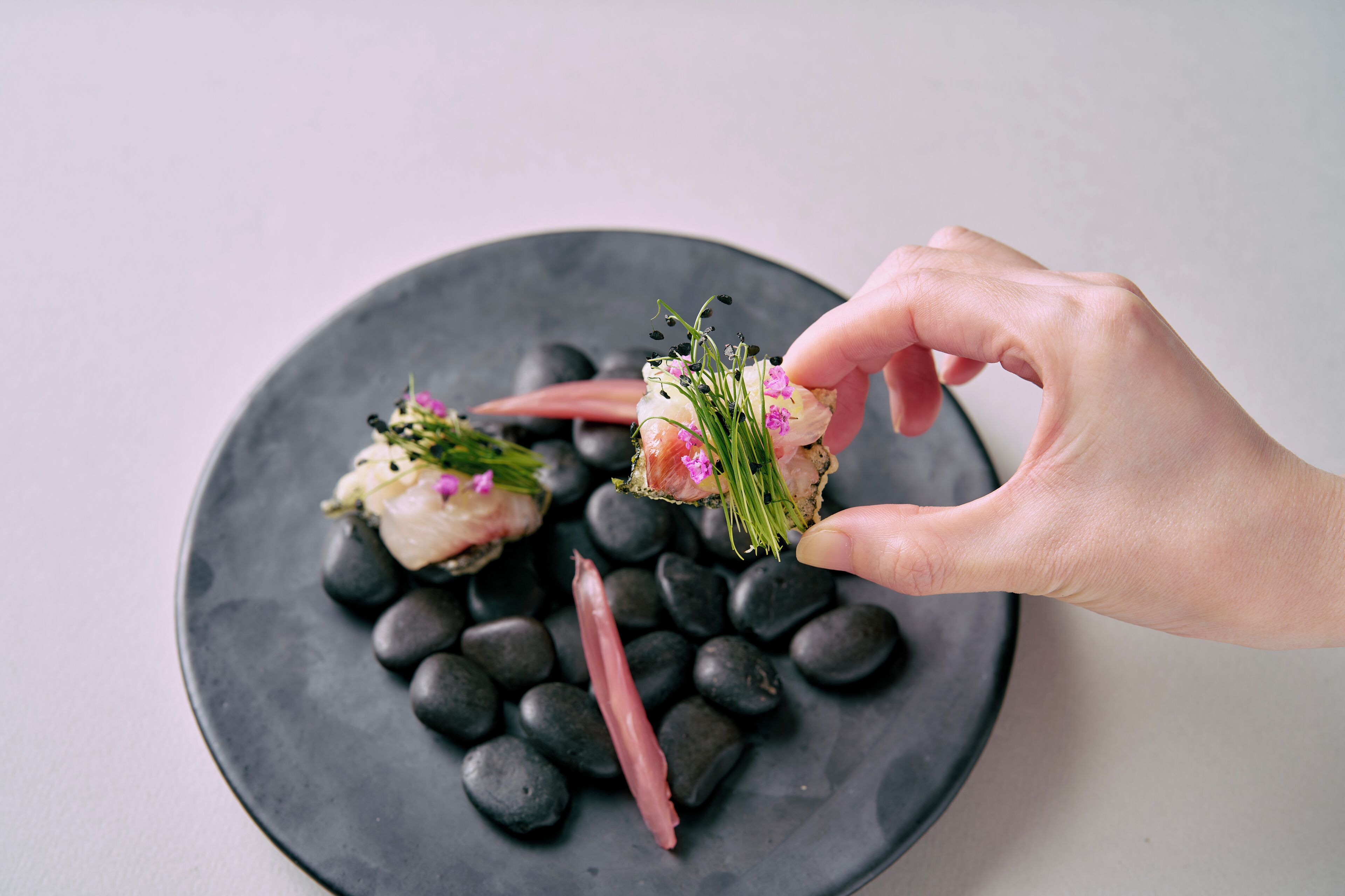 Een hand houdt een stuk sushi vast met roze vis en groene garnering op een zwart bord met zwarte stenen.
