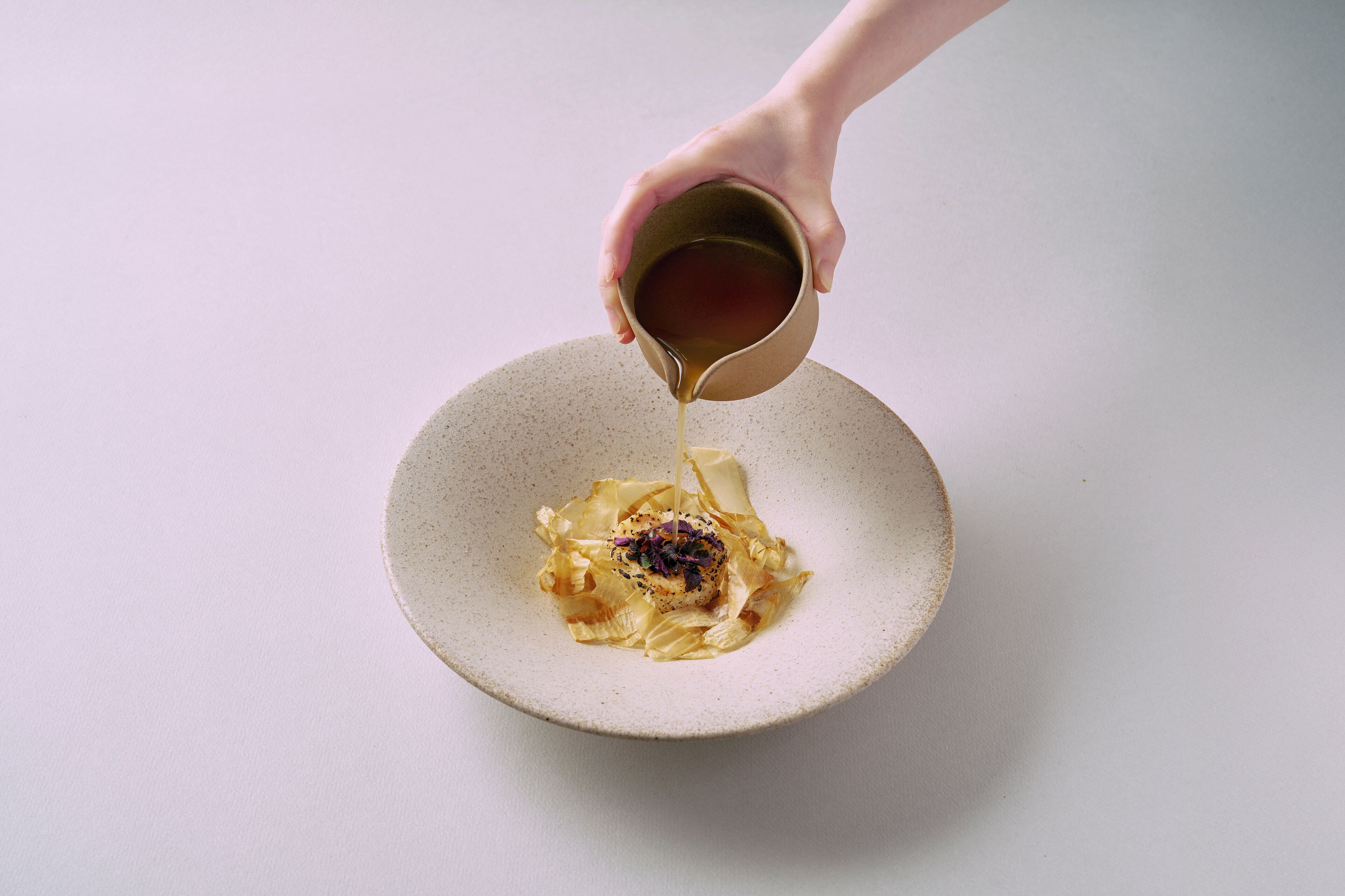 A hand pouring a liquid into a bowl with a piece of food on top.