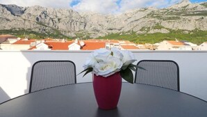 Une table sur laquelle est posé un vase rouge contenant des fleurs blanches, le tout se détachant sur un arrière-plan composé de montagnes et d’une vue sur la ville.