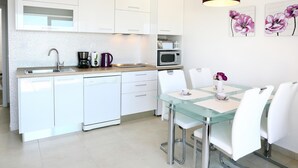 Een moderne keuken met witte kastjes, een roestvrijstalen spoelbak en een koffiezetapparaat. Er staat een eettafel met witte stoelen en een glazen blad.