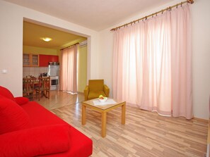 Ein Wohnzimmer mit einem roten Sofa, einem Holztisch mit einer Obstschale und einem gelben Sessel. Durch einen offenen Durchgang ist eine Küche mit roten Schränken und ein Essbereich mit Holztischen zu sehen.