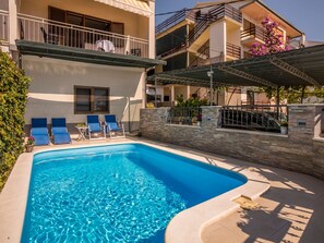 Ein Swimmingpool mit Liegestühlen, eine Steinmauer und ein mehrstöckiges Gebäude mit Balkonen.
