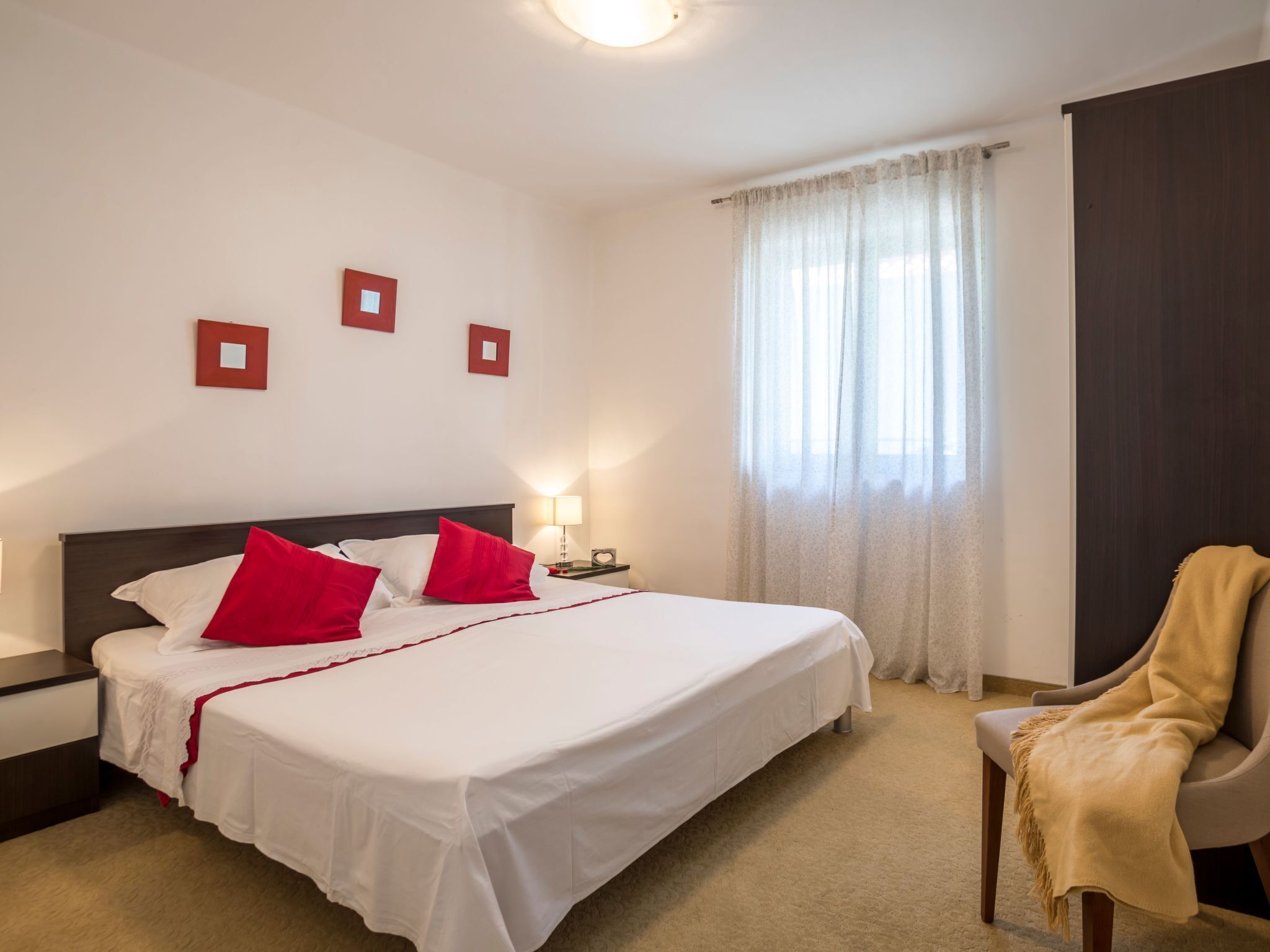 A hotel room with a large bed, a bedside table with a lamp, a chair with a blanket, and a window with curtains.