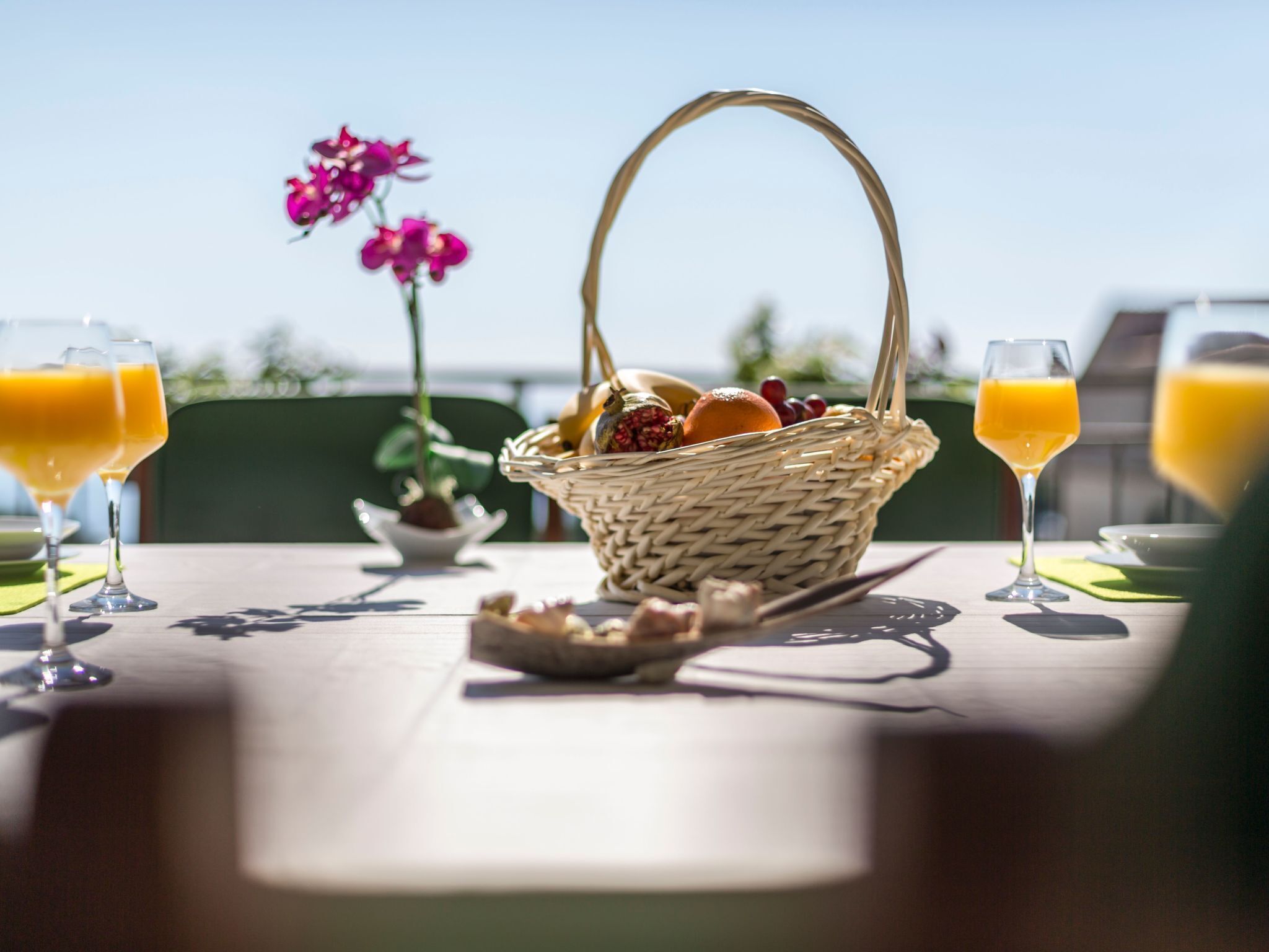 A table with a wicker basket containing fruits and a plate of bread, two glasses of orange juice, and a potted plant.