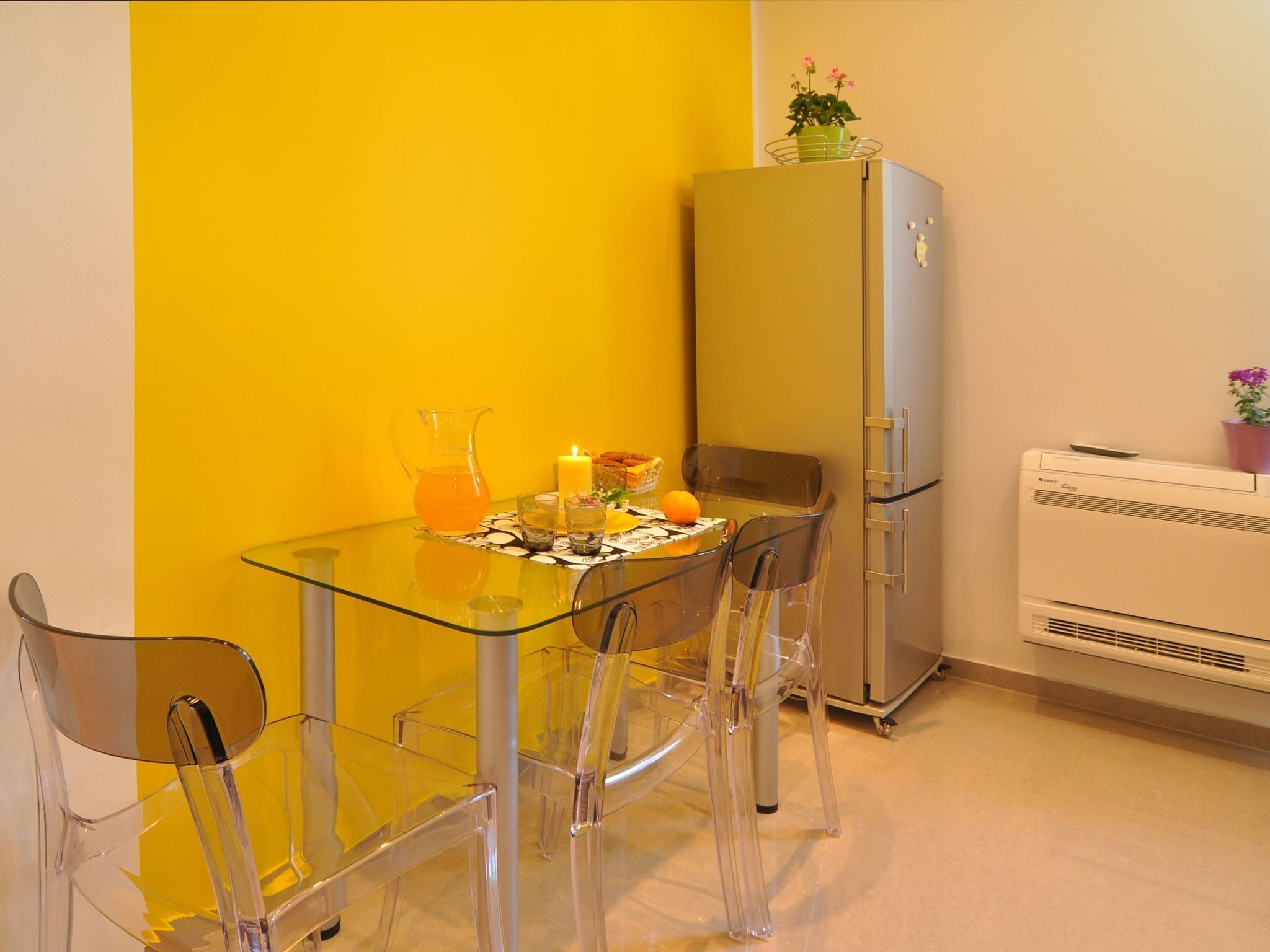 A compact kitchen with a glass table, four chairs, a refrigerator, and a small air conditioning unit.