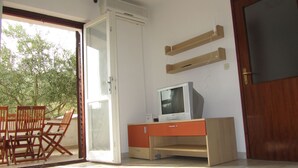 Ein Zimmer mit einer Glasschiebetür, die zu einem Außenbereich mit Tisch und Stühlen führt. Ein Fernseher steht auf einem Holzgestell, darunter befindet sich ein Schrank.