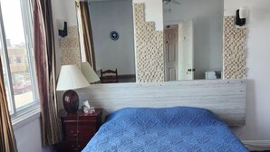 A bedroom with a large bed, a bedside table with a lamp, a mirror, and a window with curtains.
