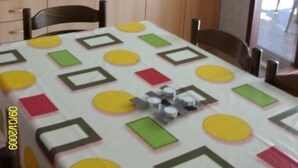 Une cuisine de taille réduite, dotée de meubles en bois, d’une cuisinière à gaz et d’un évier. On y trouve une table à manger recouverte d’une nappe colorée.