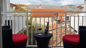 Ein Balkon mit Korbmöbeln und roten Kissen, mit Blick auf eine Berglandschaft mit Häusern mit Terrakotta-Dächern.