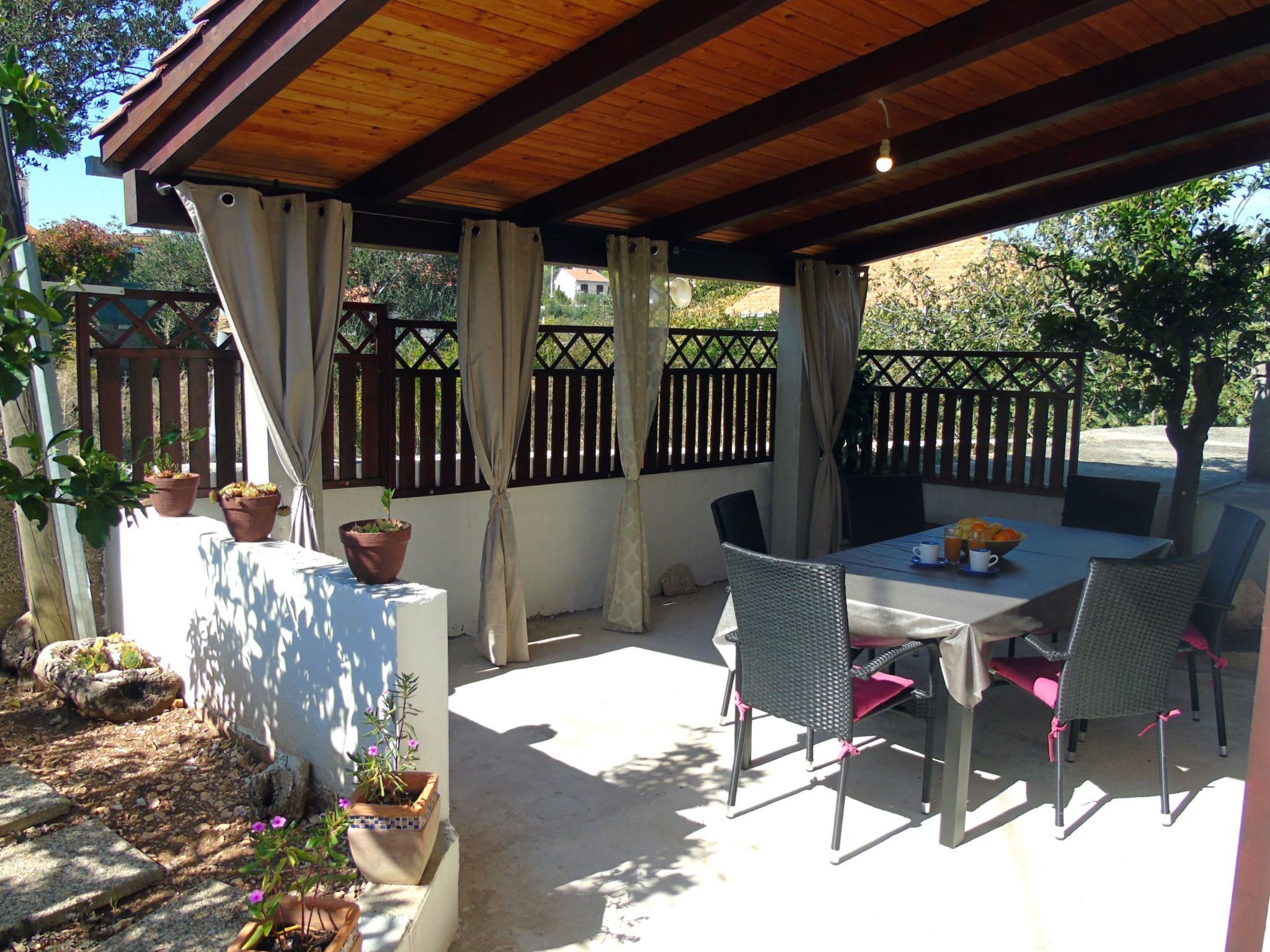 Eine Terrasse im Freien mit Tisch und Stühlen, umgeben von Kübelpflanzen und einem Holzzaun.