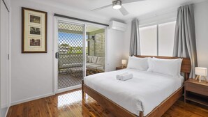 A hotel room with a wooden floor, a bed with white linens, a ceiling fan, a window with curtains, and a door leading to a balcony. - Bright 3-Bed Home in Leafy Annerley (Annerley)