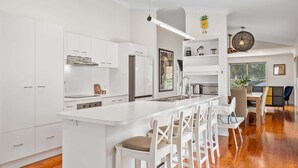 A modern kitchen with white cabinetry, a central island with bar stools, and a dining area with a wooden table and chairs. - Bright 3-Bed Home in Leafy Annerley (Annerley)