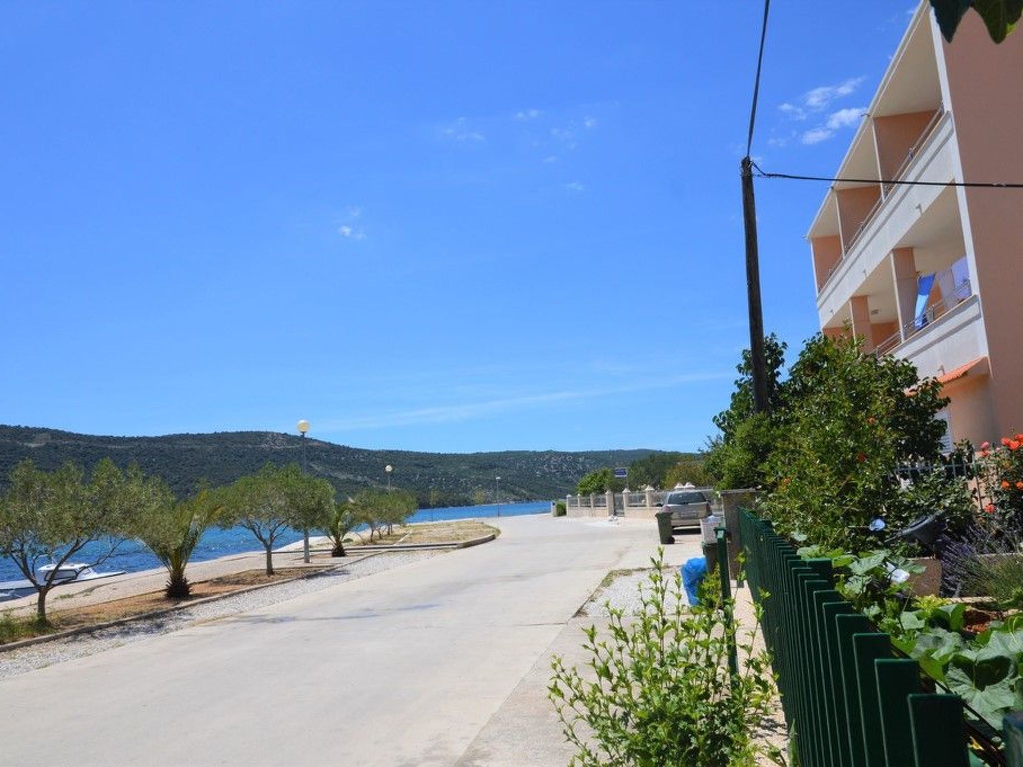 Uma estrada costeira com um edifício à direita, céu azul límpido e uma paisagem acidentada ao fundo.