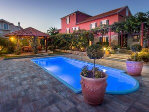 Une piscine agrémentée d’une pergola et d’un espace salon extérieur, le tout entouré d’un jardin bien entretenu et d’une grande maison à la façade rouge.