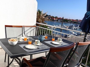 Une table de balcon dressée pour le petit-déjeuner, avec des assiettes, des verres et un bol, donnant sur une marina où sont amarrés des bateaux, sous un ciel dégagé.