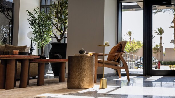 Un vestíbulo amplio con diseño moderno, que incluye una zona de estar con una mesa y sillas de madera, una planta grande en maceta y vistas al exterior a través de amplias ventanas.