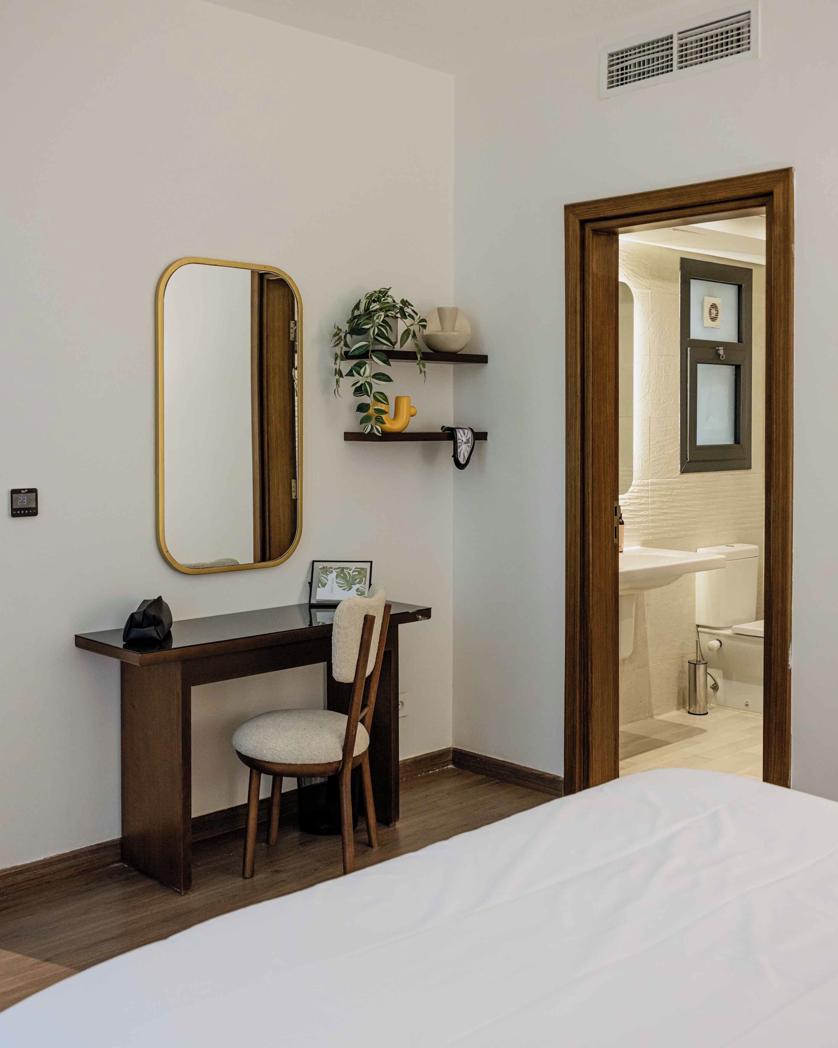 A bedroom with a bed, a desk with a chair, a mirror, and a bathroom visible through an open door.