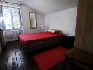 A small, single-bed room with a wooden floor, a red bedspread, a white fan, and a window with floral curtains. - Schönes Haus mit privatem Garten by Interhome (Zadar)