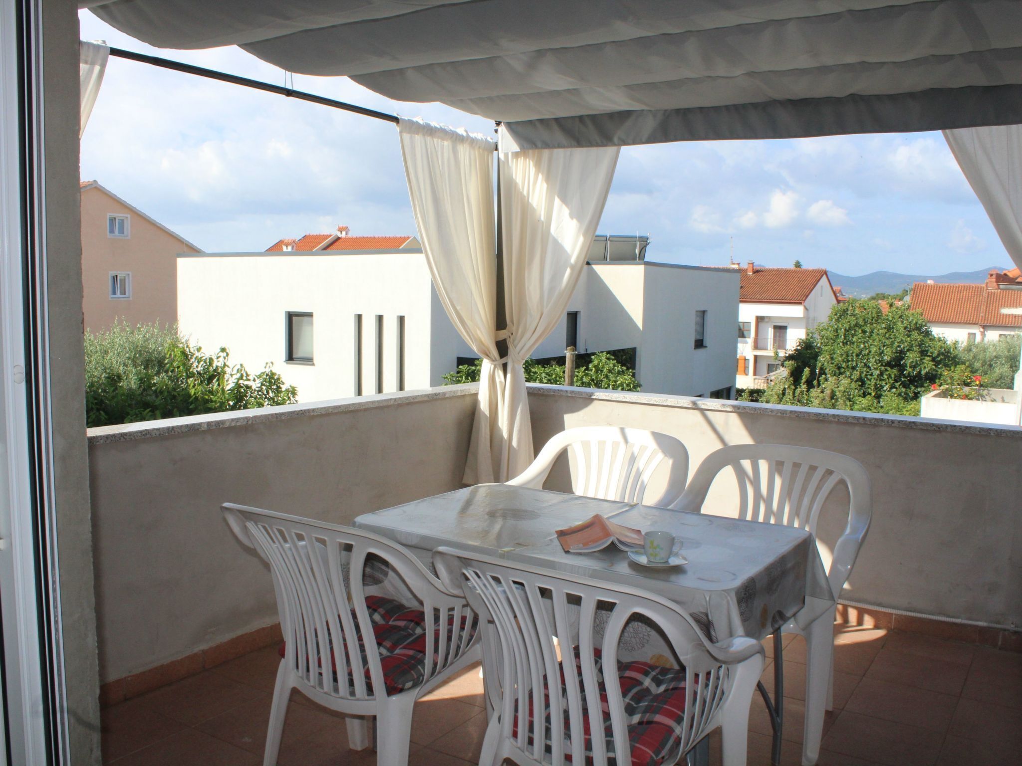 Un balcon avec une table et des chaises, un rideau blanc et une vue sur des immeubles et des arbres.