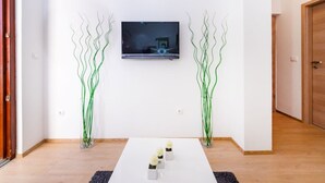 Ein modernes Wohnzimmer mit einem an der Wand befestigten Flachbildfernseher, einem weißen Couchtisch mit Dekoration und zwei hohen, grünen Pflanzenskulpturen.
