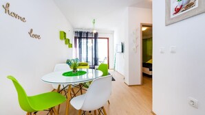 Ein moderner Essbereich mit einem Glastisch, grünen und weißen Stühlen und Blick in einen Raum mit Bett und Fernseher.