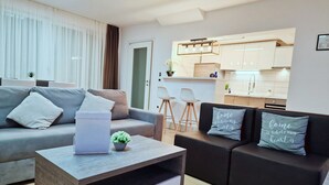 Un salon moderne avec un canapé gris, un canapé noir et une table basse en bois. On y trouve également un coin cuisine avec des meubles blancs et un tabouret de bar.