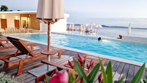 Un espace aménagé au bord de la piscine, avec des chaises longues en bois et un grand parasol, offrant une vue sur une piscine aux eaux cristallines et sur la mer.