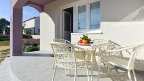 Eine überdachte Terrasse mit Tisch und Stühlen, ein Fenster mit Jalousien und eine Obstschale auf dem Tisch.