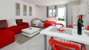 Ein modernes Wohnzimmer mit einem roten Sofa, einem weißen Couchtisch und einem Essbereich mit roten Stühlen und einem weißen Tisch. Im Hintergrund ist ein Bett mit einer rot-weißen Tagesdecke zu sehen.