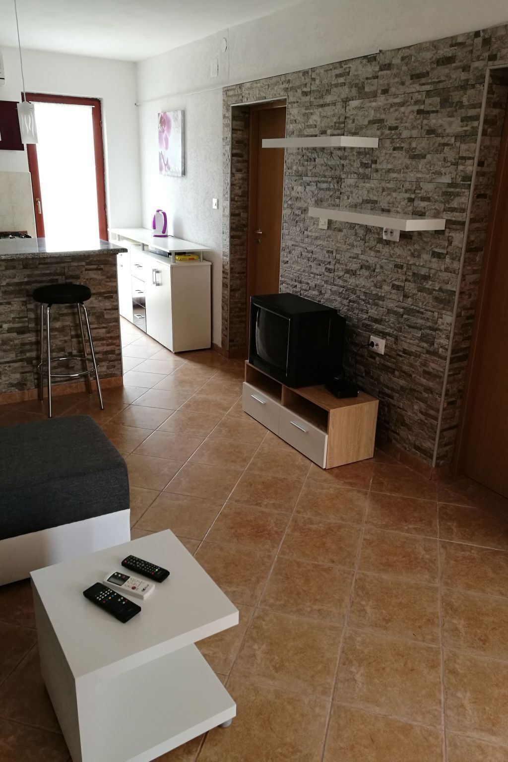 Un espacio habitable compacto con área de cocina, un televisor sobre un soporte de madera y una mesa de centro blanca con controles remotos.