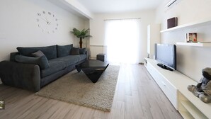 Una sala de estar moderna con un sofá gris, una mesa de centro negra, un televisor sobre un soporte blanco, un reloj de pared y una planta.