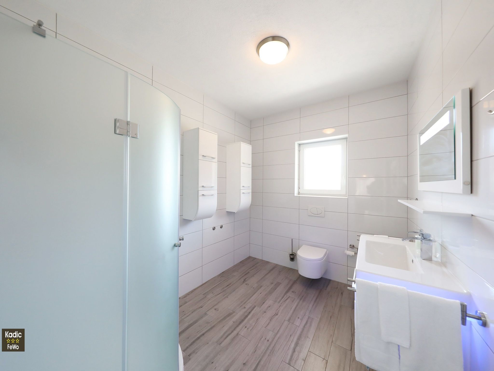 A modern bathroom with white tiles, a wooden floor, a toilet, a sink, and a window.