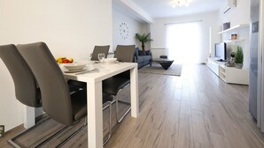 Una sala de estar moderna con comedor, que incluye una mesa blanca, sillas grises, un sofá y un televisor.