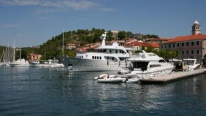 A marina with several yachts docked, a prominent building with a red roof in the background, and a clear blue sky.
