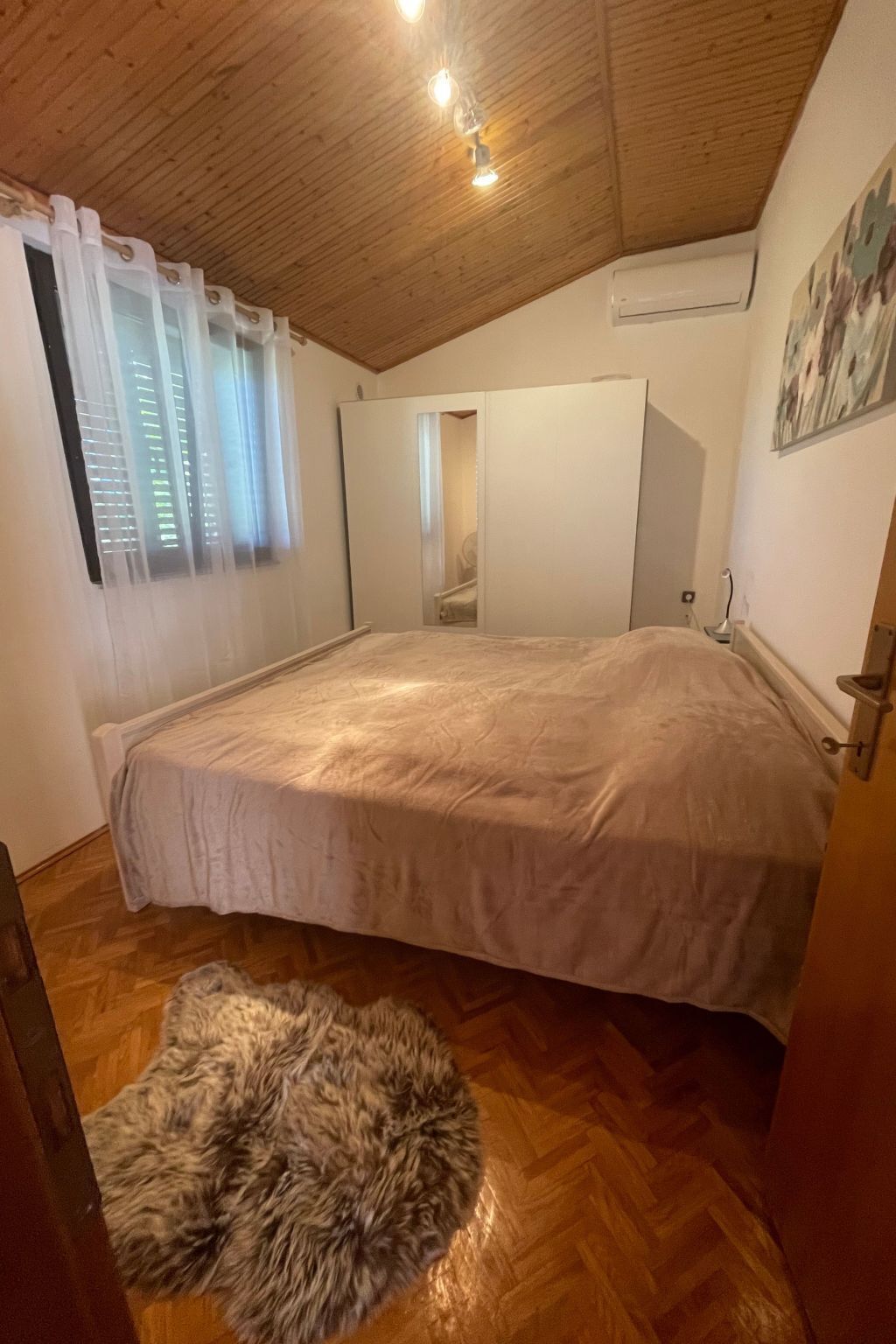 A bedroom with a large bed, wooden flooring, a fur rug, a wardrobe, and a ceiling light fixture.