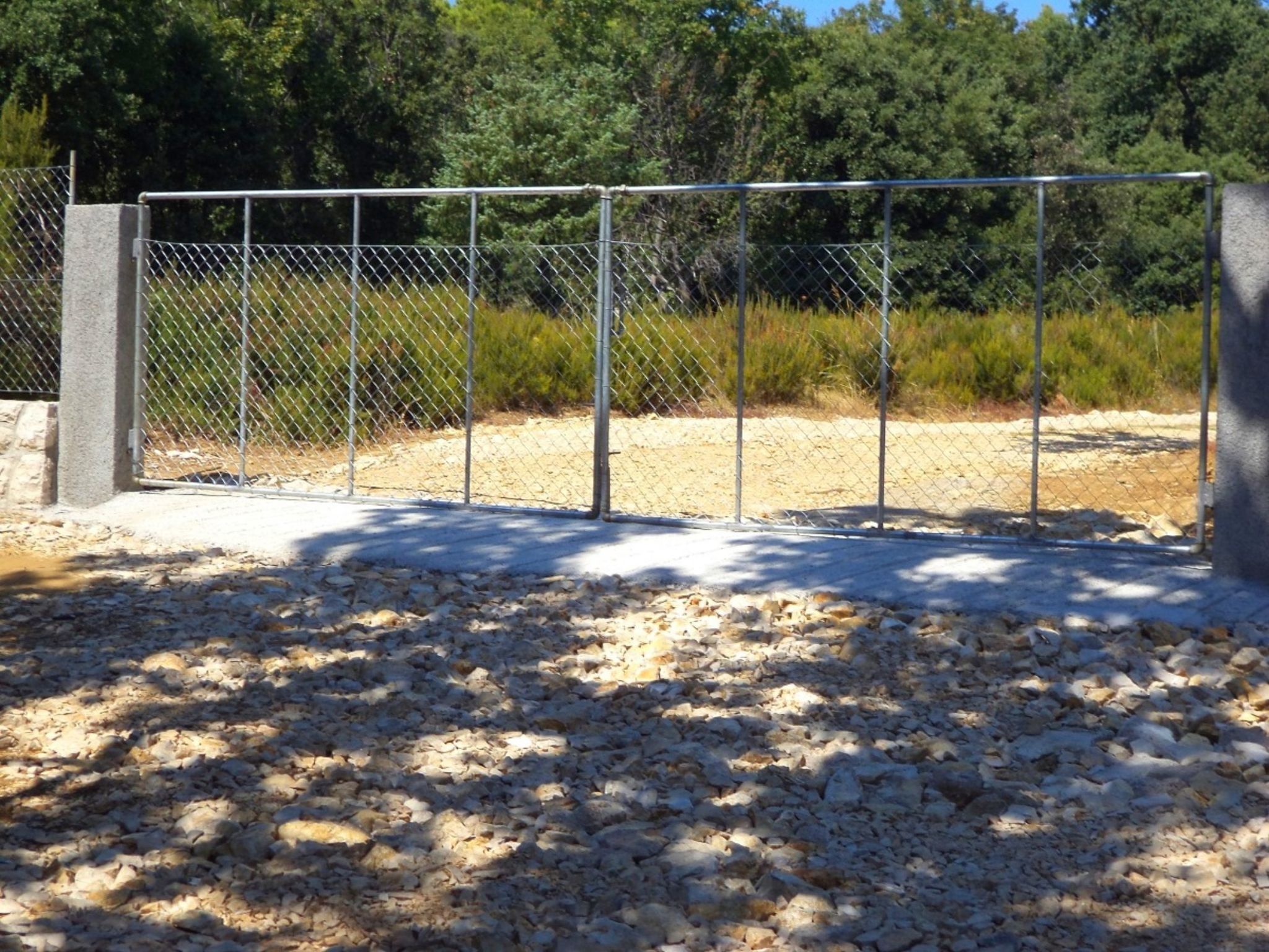 A chain-link fence with a concrete base, set against a backdrop of trees and a gravel path.