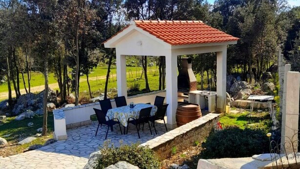 A covered outdoor seating area with a table and chairs, surrounded by a stone patio and a garden with trees and rocks. - in Kampor mit Privatparkplatz by Interhome (Rab/Kampor)