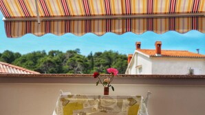 A balcony with a striped awning, a small table with a vase of flowers, and a view of buildings and trees. - Appartement in Palit mit Privatem Balkon by Interhome (Rab/Rab)