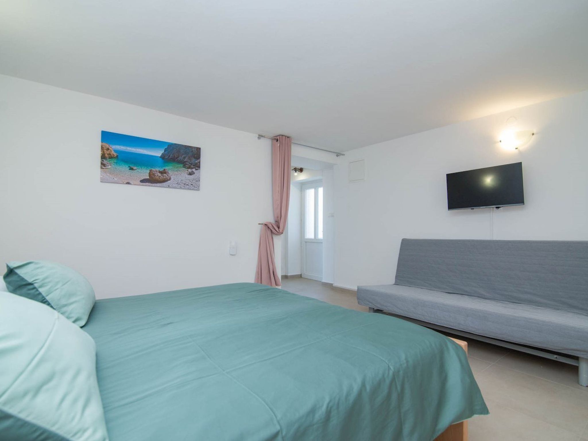 Une chambre à coucher avec un lit, un canapé gris, un téléviseur fixé au mur et un tableau représentant une scène de plage.
