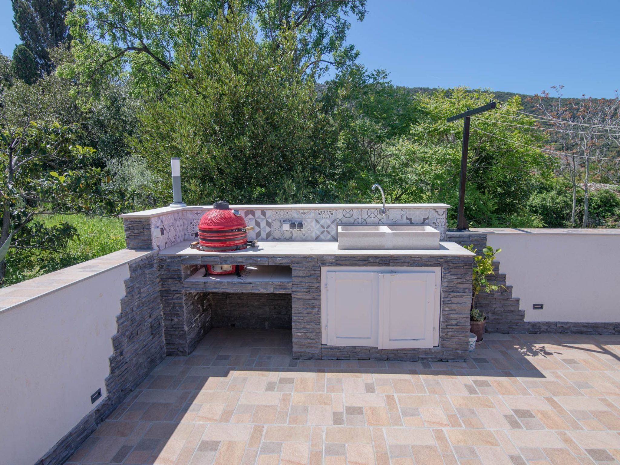 Une cuisine extérieure dotée d’un plan de travail en pierre, d’un évier encastré et d’un barbecue rouge.