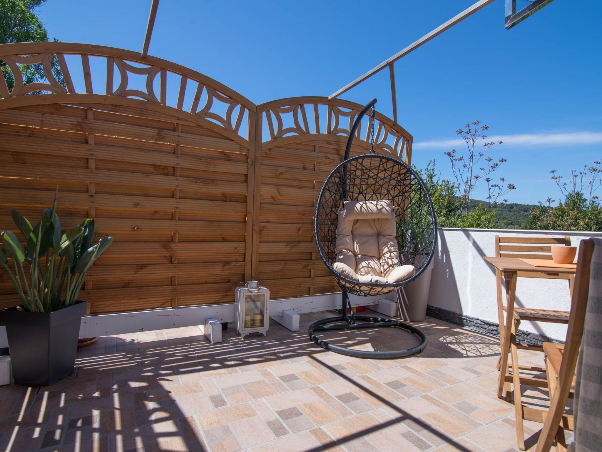 A wooden pergola with a hanging chair, a potted plant, and a table with a bowl.
